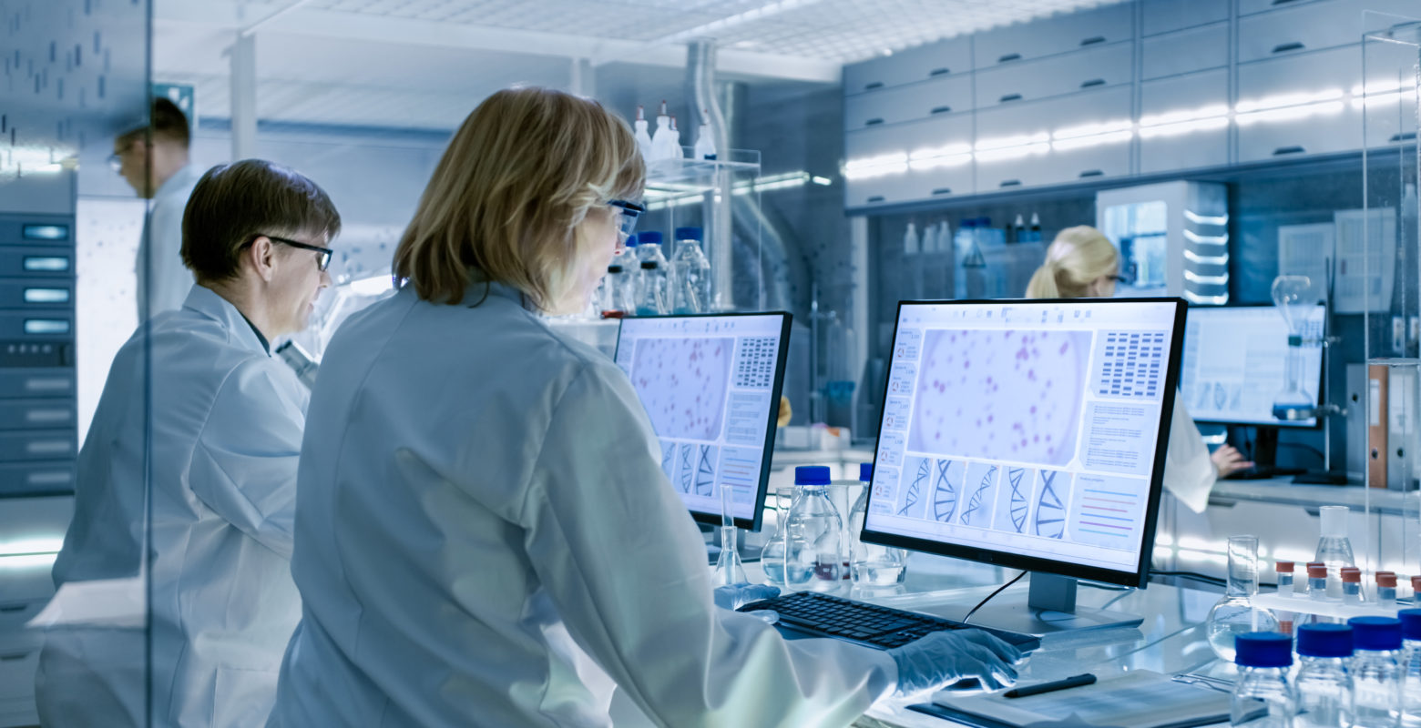 Female and Male Scientists Working on their Computers In Big Modern Laboratory. Various Shelves with Beakers, Chemicals and Different Technical Equipment is Visible.